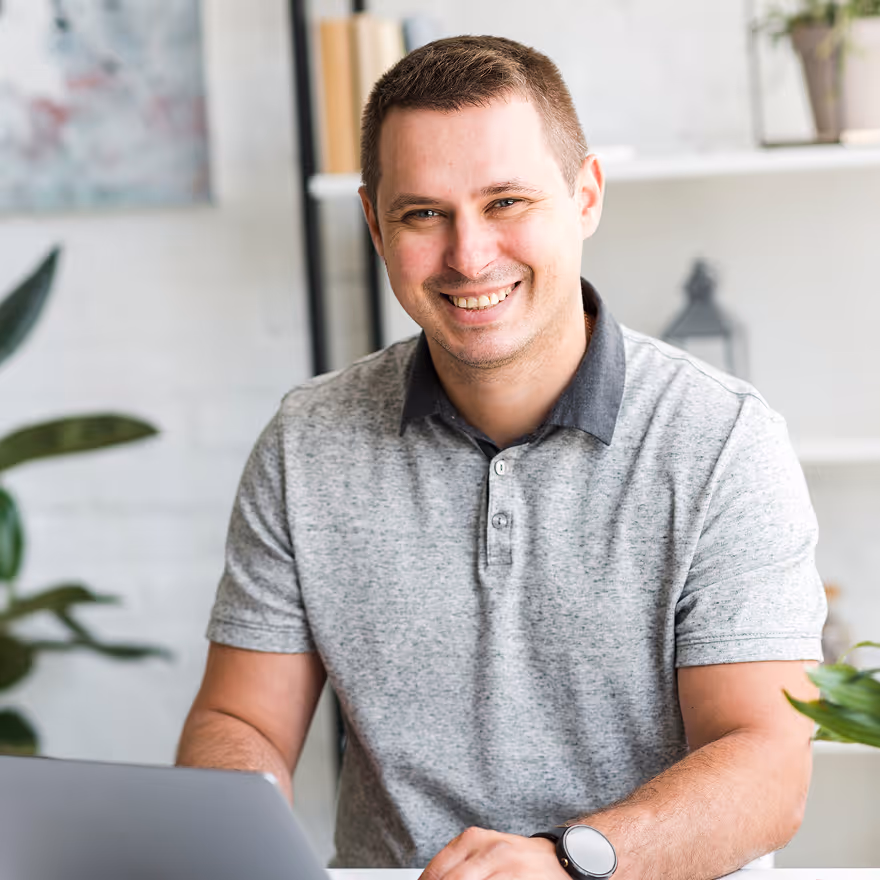 Smiling man wearing a gray polo shirt working on a laptop in a bright room with plants and shelves in the background.