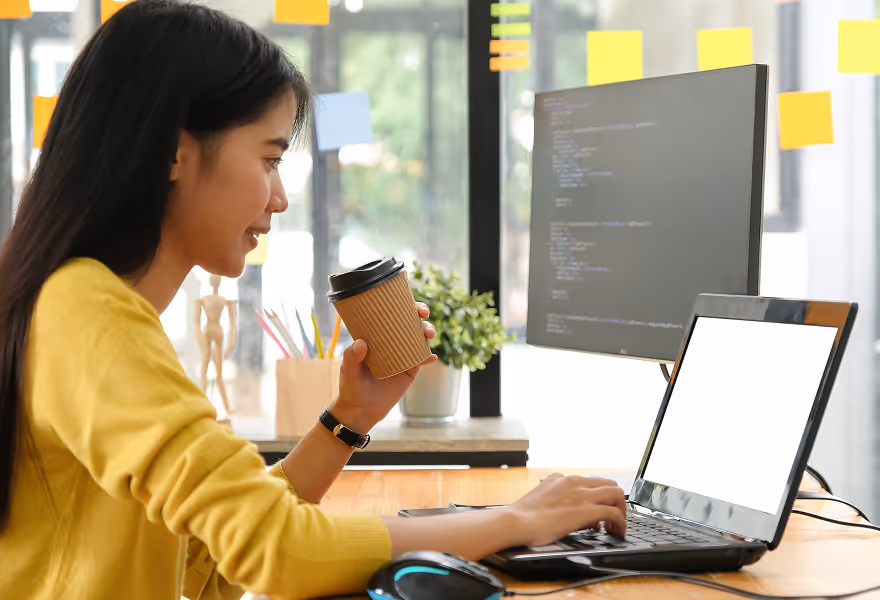 Woman in yellow sweater holding a coffee cup and typing on a laptop with a code editor displayed on a monitor in the background.