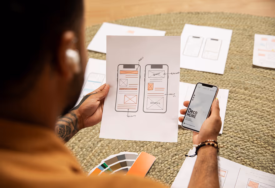 Person holding a paper with hand-drawn mobile app wireframes and a smartphone displaying a Deco Home app interface, surrounded by design sketches and color swatches on a woven surface.