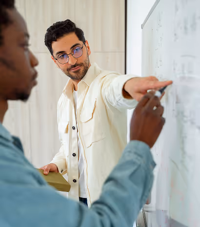 Man wearing glasses pointing at a whiteboard with text while another man writes on it.