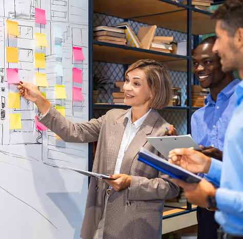 Businesswoman pointing to sticky notes on a whiteboard while discussing with two male colleagues in an office.