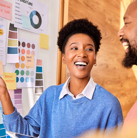 Smiling woman in a blue sweater discussing colorful charts and sticky notes with a man in an office setting.
