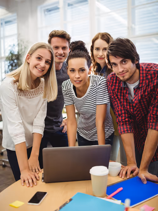 Five young adults gathered around a laptop in a bright office, smiling and leaning on a table with coffee cups and notebooks.
