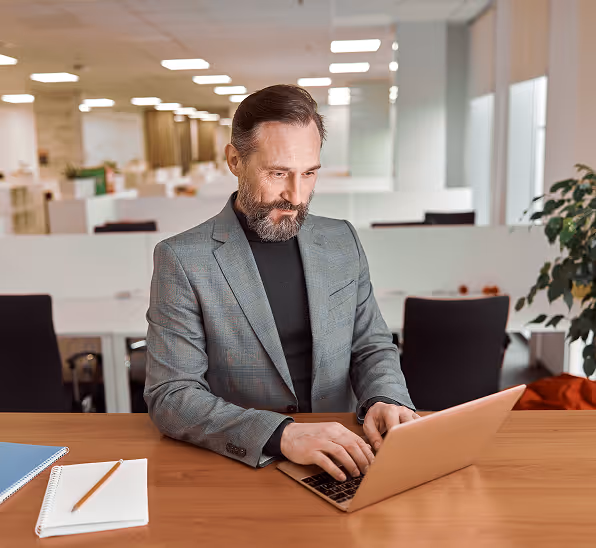 Bearded man in a gray blazer working on a laptop at a wooden desk in a modern office.