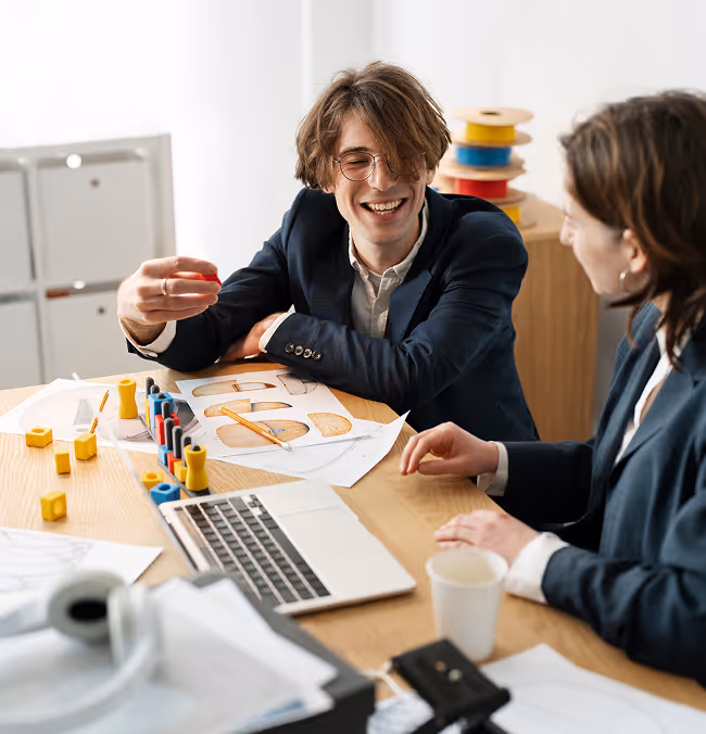 Two people in business attire discussing colorful geometric shapes and drawings at a desk with a laptop.