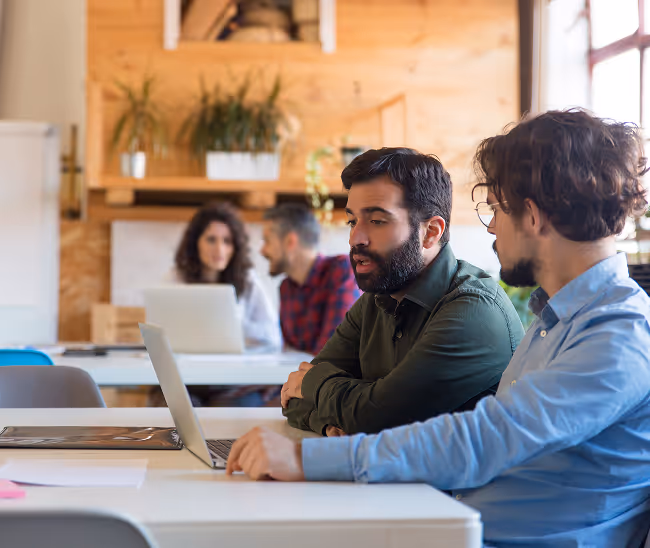 Two men working together on a laptop in a modern office with two colleagues in the background.