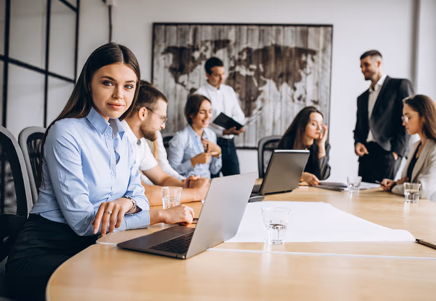 A diverse group of businesspeople in a meeting room with laptops and notebooks, one woman in a blue shirt looking at the camera.