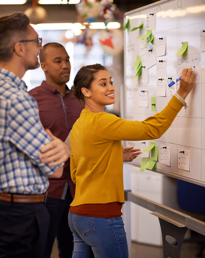Three colleagues collaborate as a woman in a yellow sweater writes on a whiteboard covered with notes and green sticky notes.
