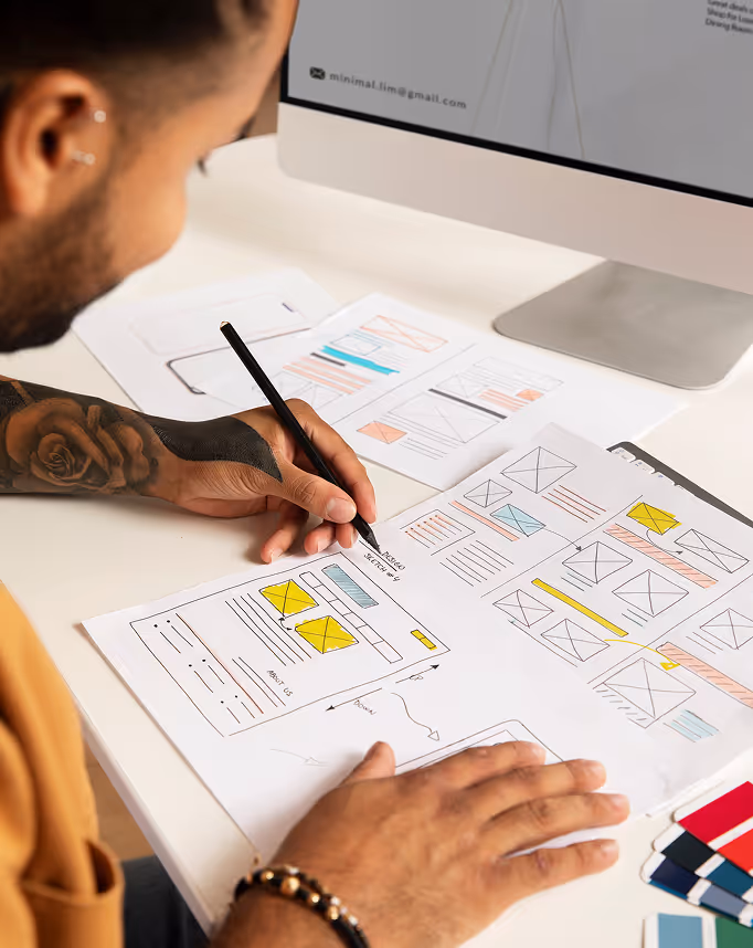 Person sketching a website wireframe with colored highlights on paper at a desk near a computer monitor.