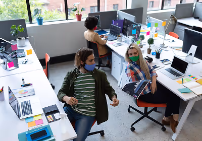 Two coworkers wearing face masks sharing a printed document while seated at desks in an office.