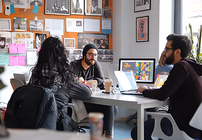 Three people engaged in a discussion around a table with laptops and coffee cups in a creative office space.
