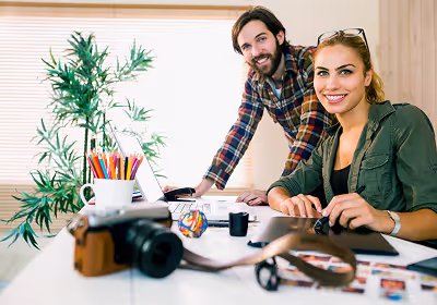 Two smiling young professionals working together at a desk with a camera, colored pencils, and documents.