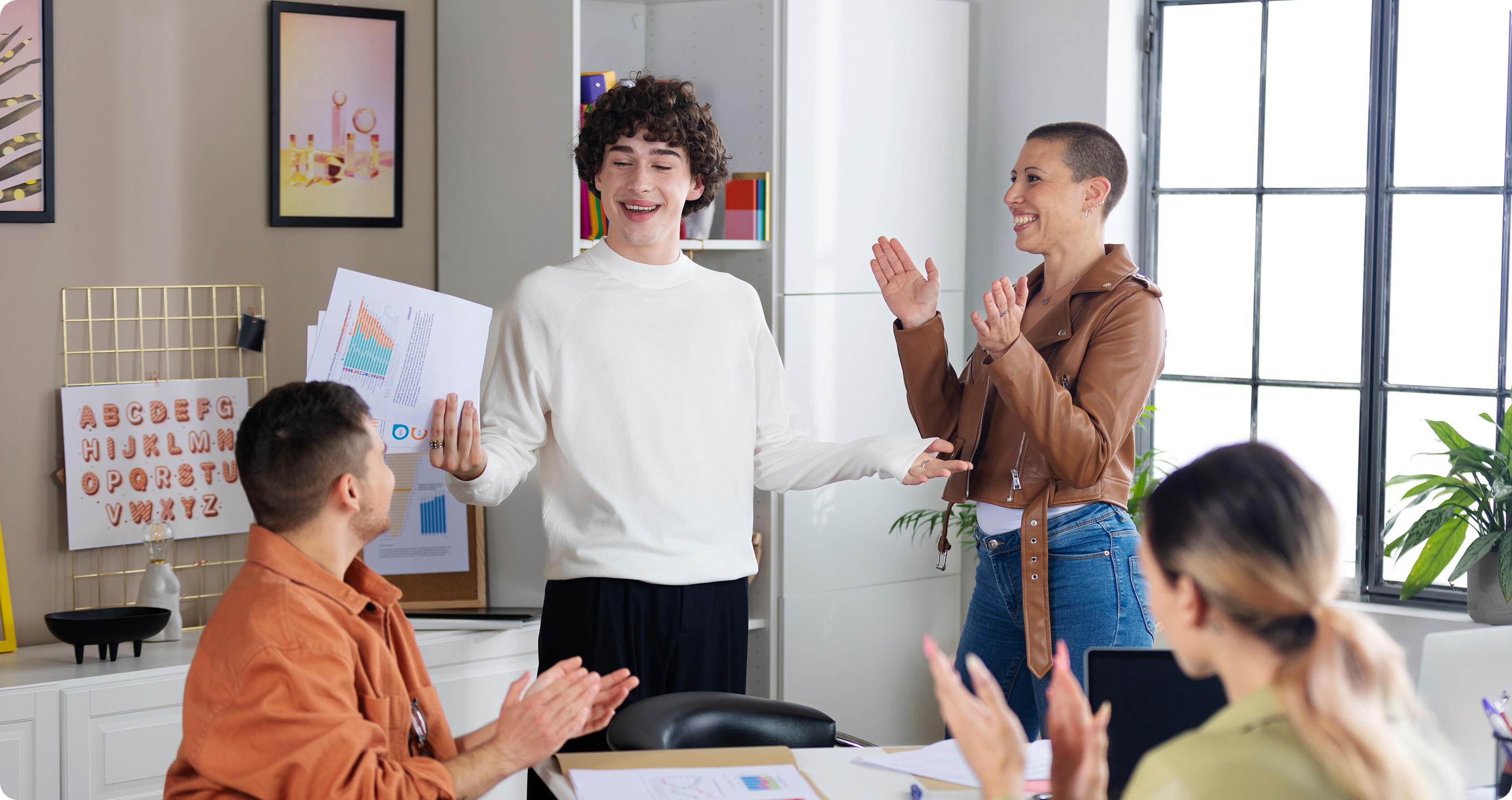 Four colleagues in an office celebrating a presentation success. A young man in a white sweater holds charts and smiles while standing. Two coworkers, one in a brown leather jacket and another seated, are clapping and smiling, showing appreciation and encouragement.