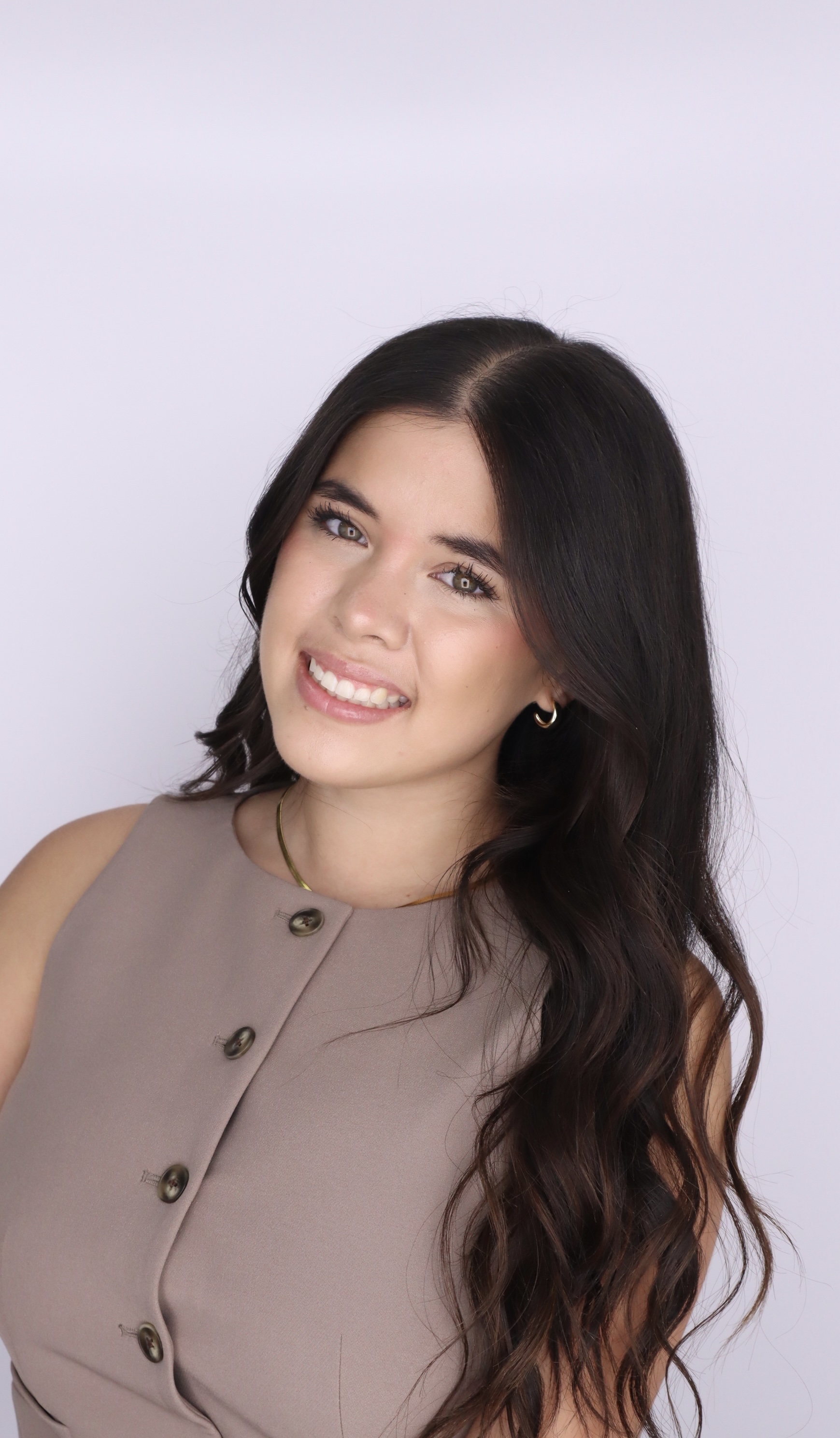Smiling woman with long wavy dark hair wearing a sleeveless taupe buttoned top and small hoop earrings.