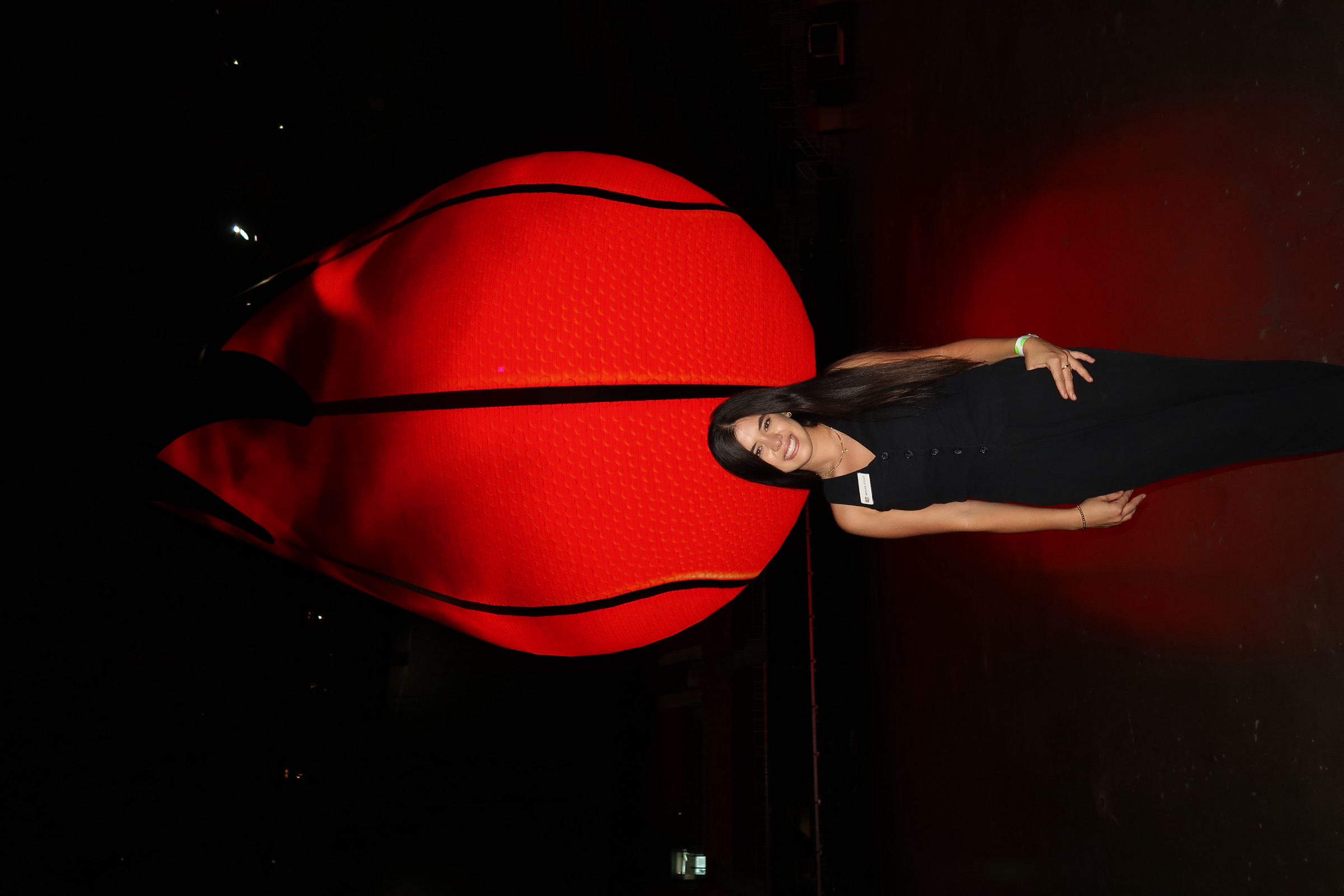 Smiling woman in black outfit standing in front of a large illuminated red basketball decoration at night.