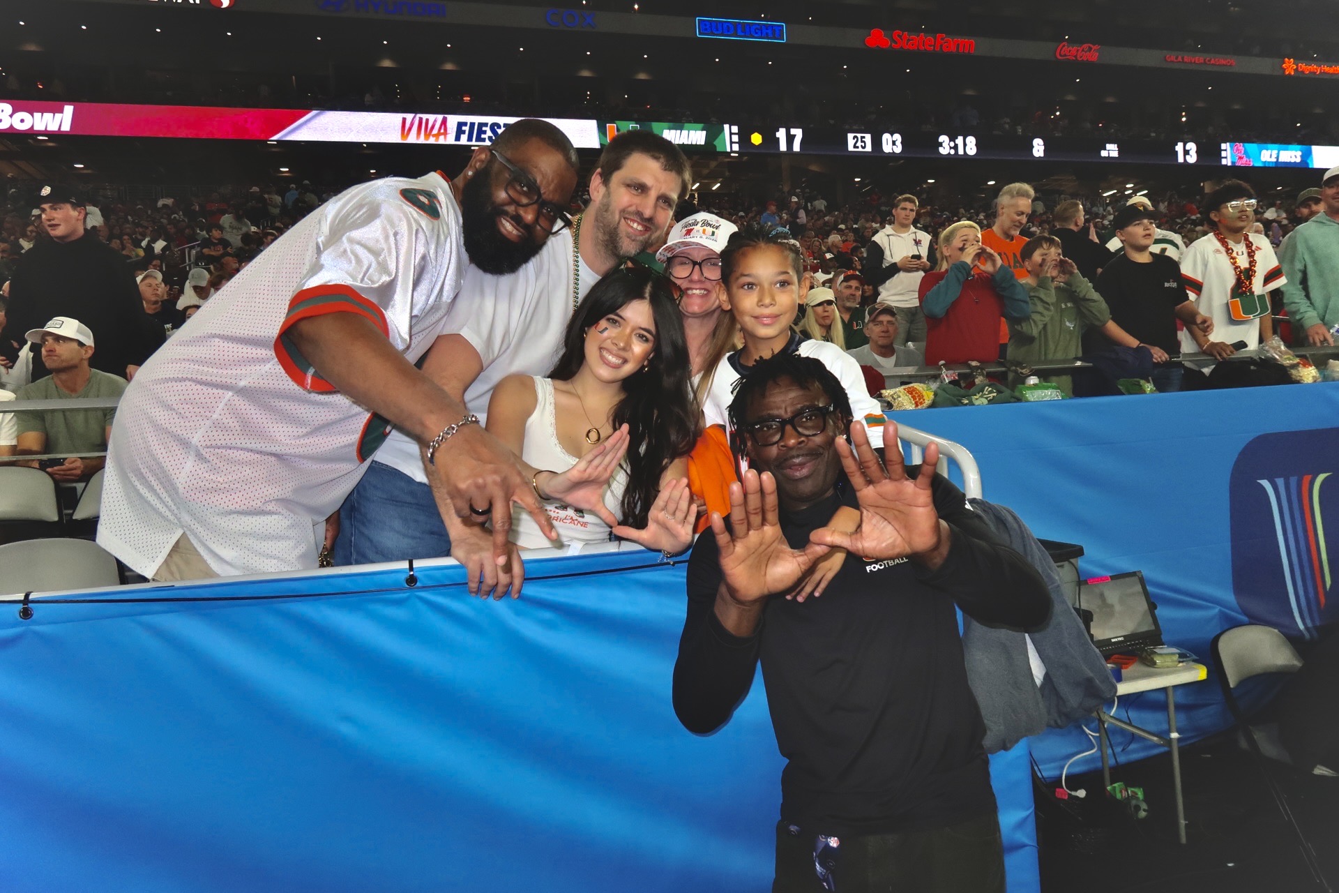 Group of diverse football fans smiling and posing with hands forming a 'U' sign at a stadium during a game.