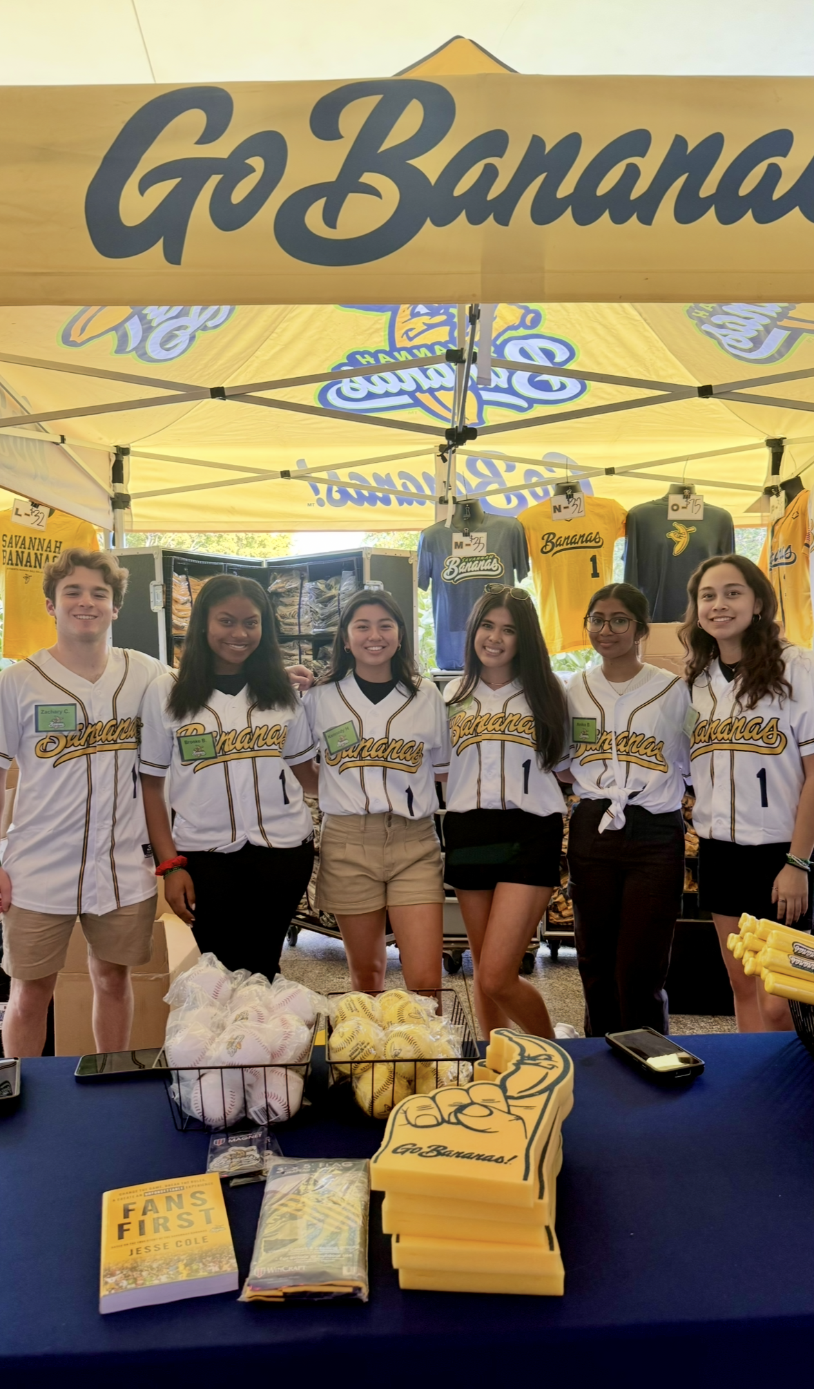 Six young adults wearing white Go Bananas jerseys standing behind a table with baseballs, softballs, foam fingers, and merchandise under a yellow Go Bananas tent.