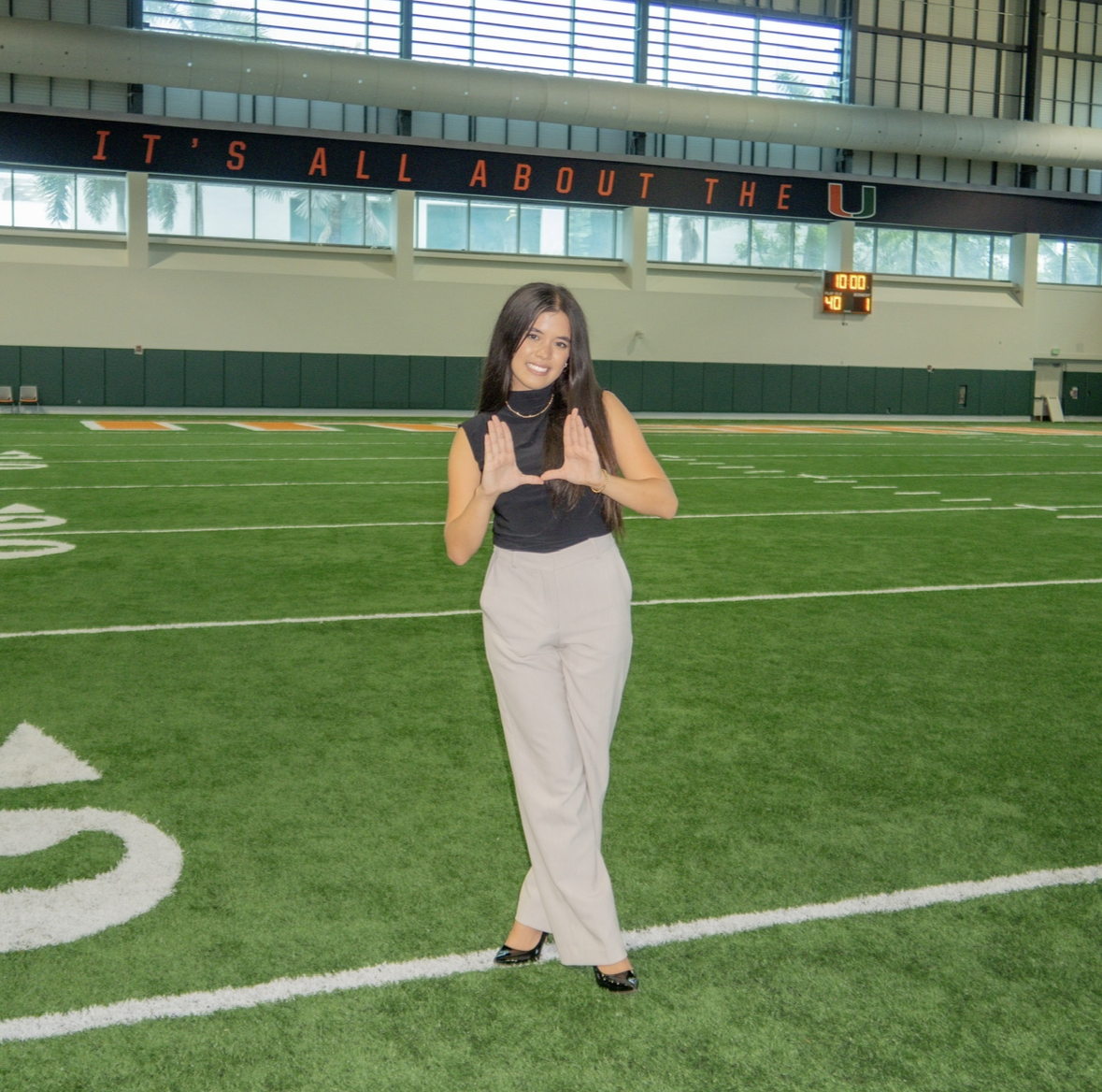 Young woman standing on an indoor football field making a 'U' hand gesture with both hands.