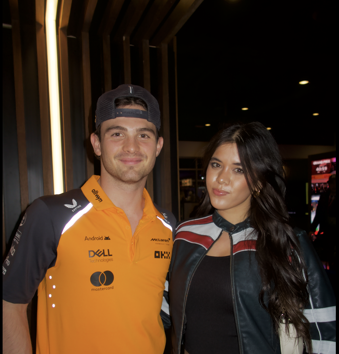 A man wearing a McLaren racing team shirt and a backwards cap stands next to a woman in a black and red leather jacket indoors at night.