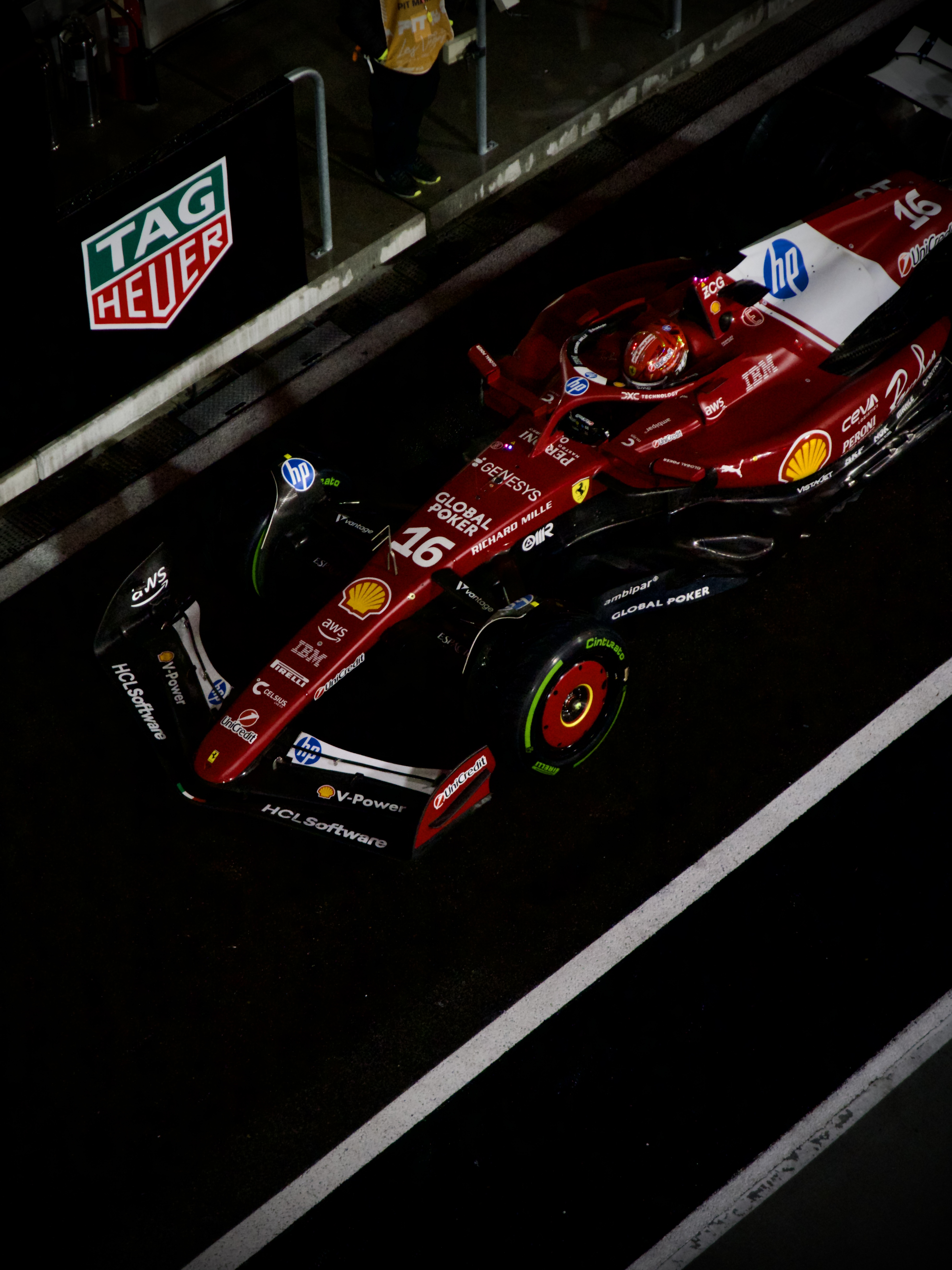 Top-down view of a red Formula 1 Ferrari race car number 16 in the pit lane at night, surrounded by sponsor logos including Shell and HP.