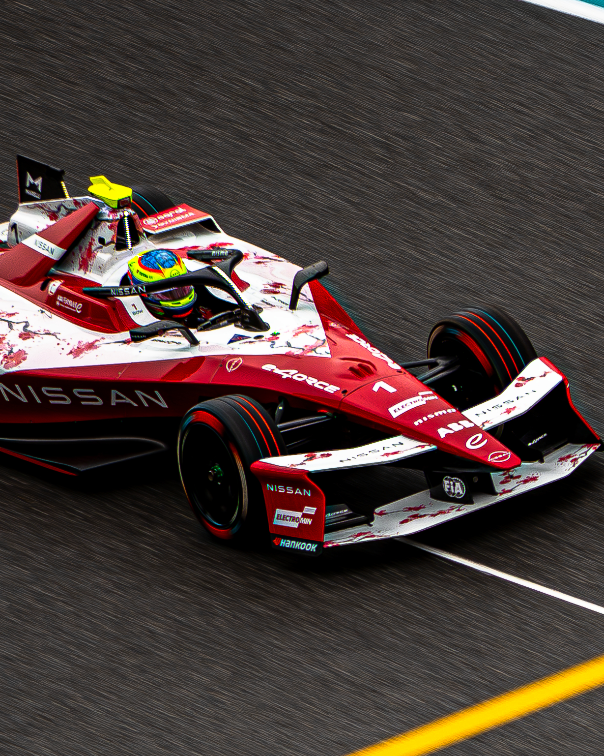 Red and white Nissan Formula E race car with driver wearing a blue and yellow helmet on a racetrack.