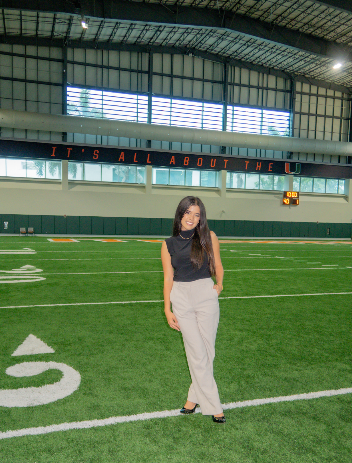 Young woman in black top and beige pants standing on an indoor football field with 'It's all about the U' sign in the background.