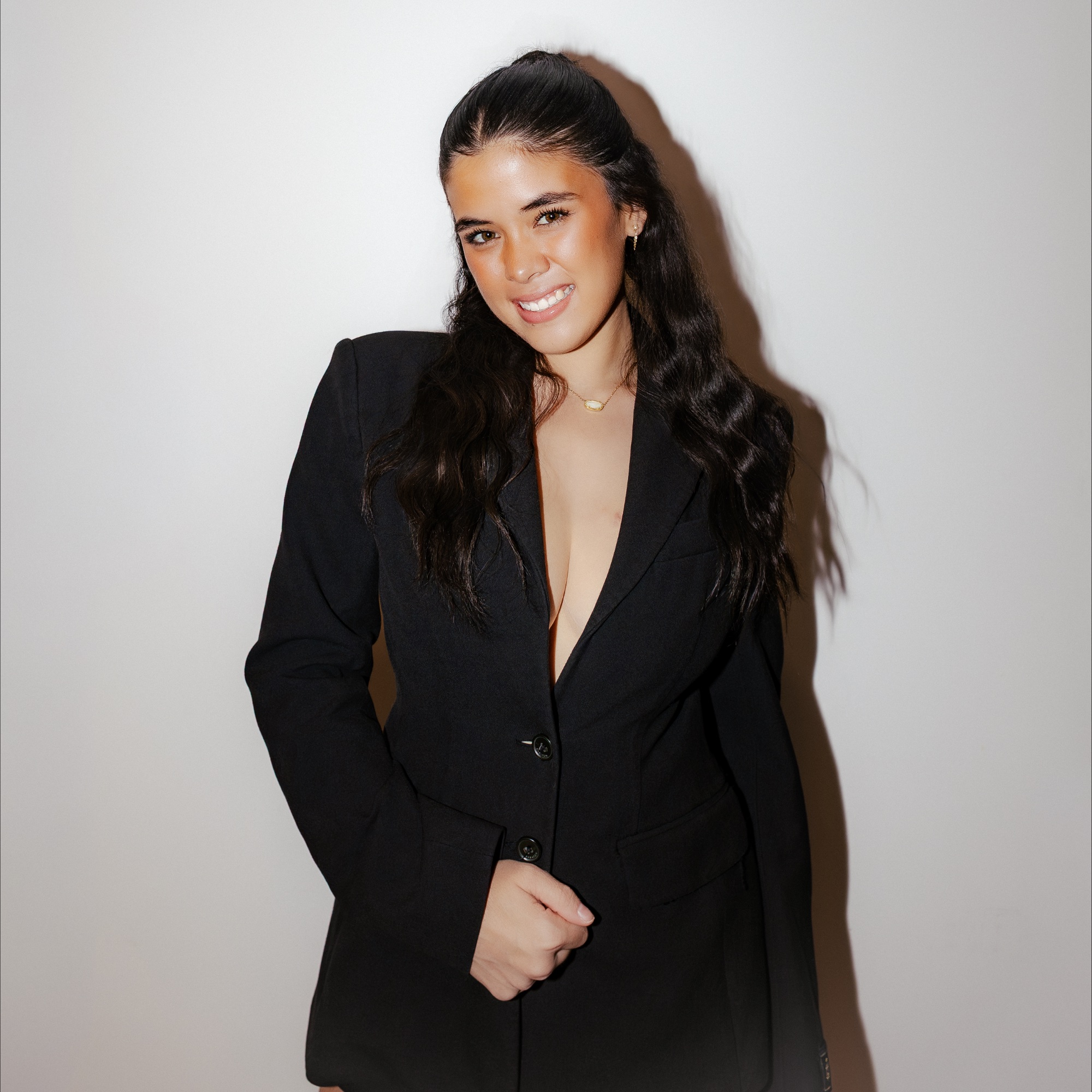 Smiling woman with long dark hair wearing a deep V black blazer against a plain background.