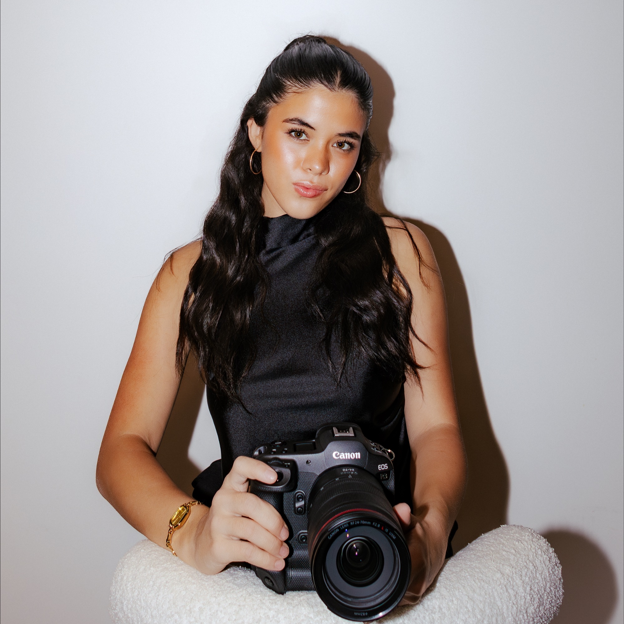 Young woman with long dark hair holding a Canon EOS camera, seated against a plain background.