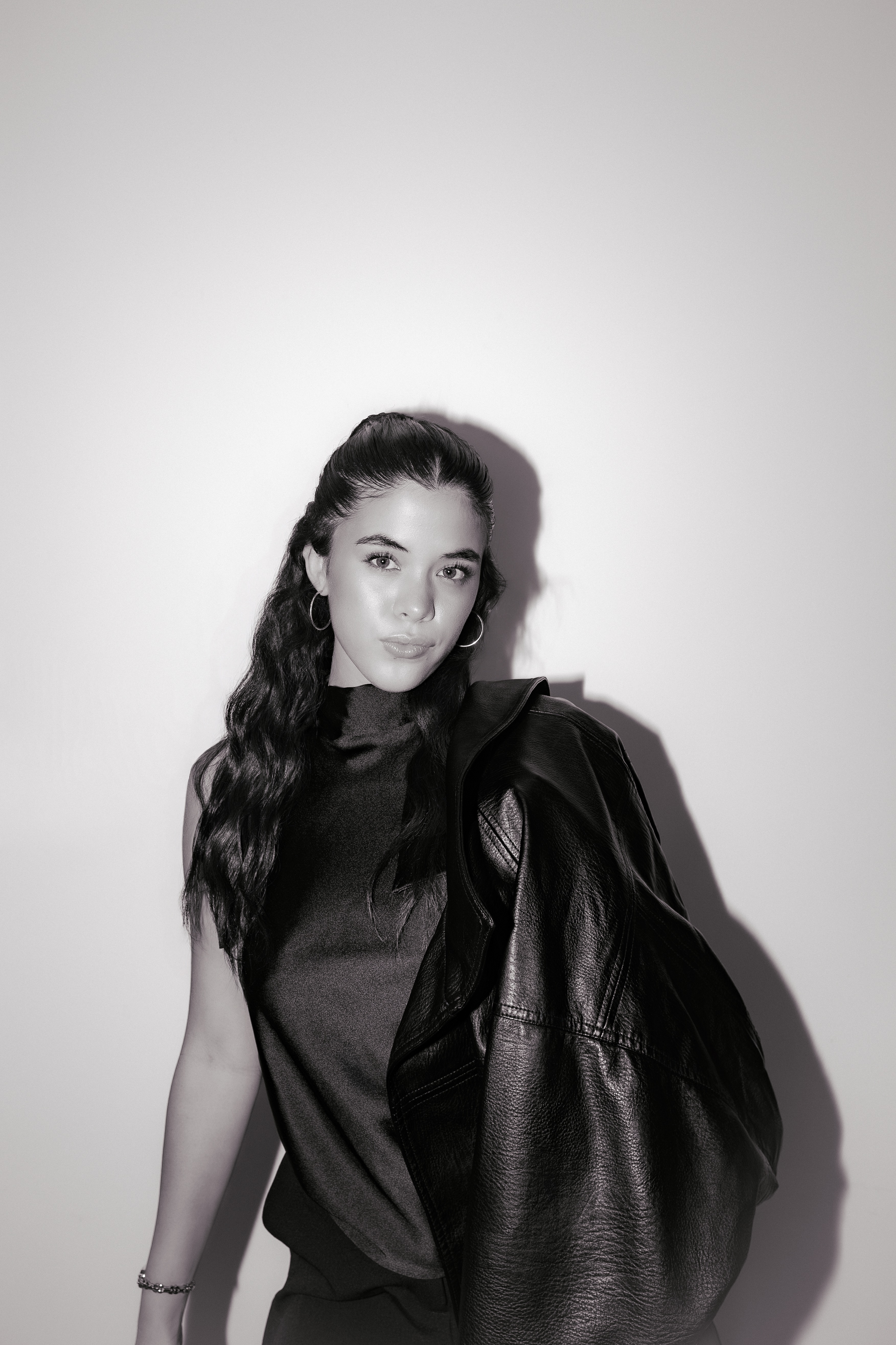 Young woman with long dark hair wearing hoop earrings and a leather jacket over one shoulder.