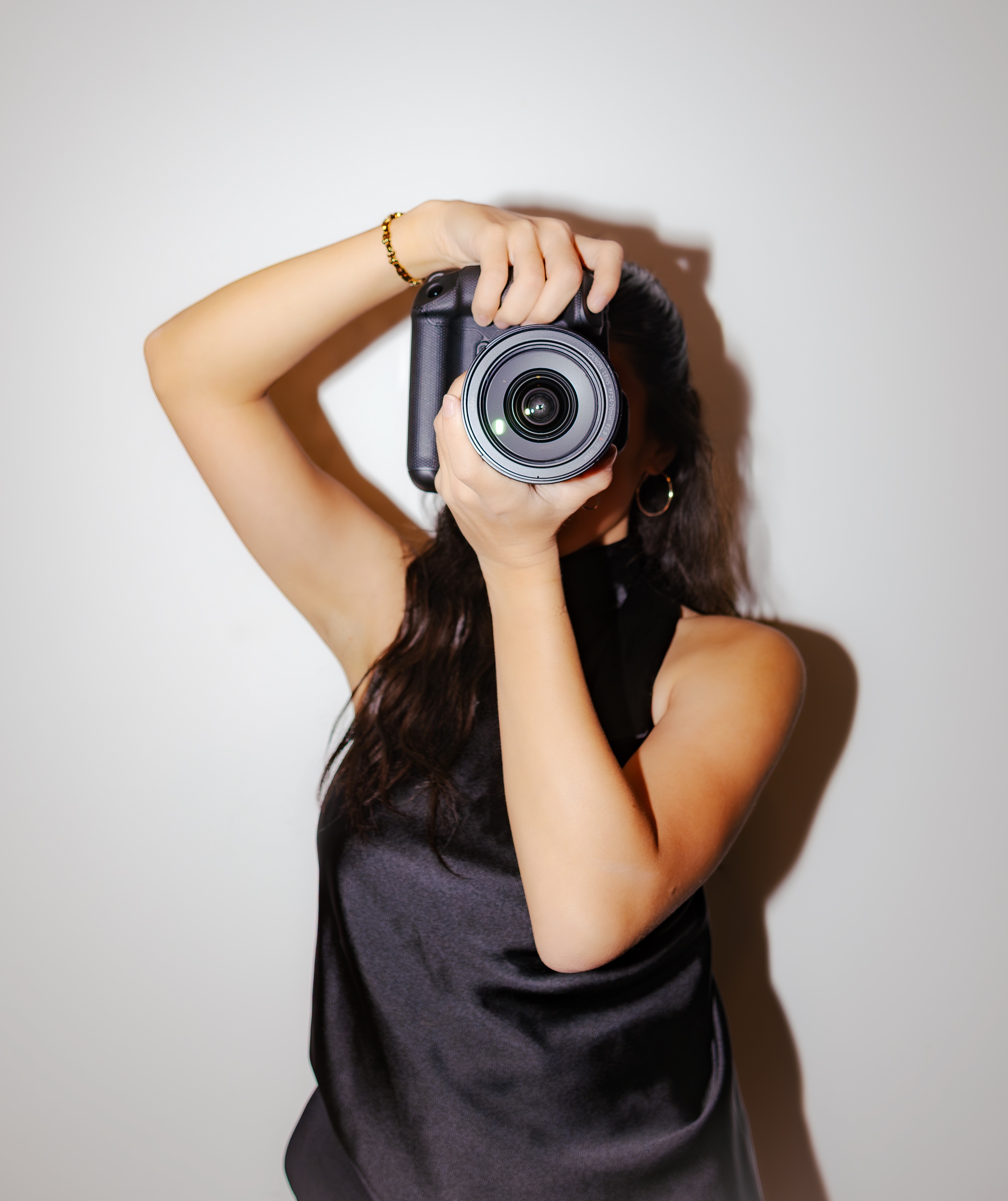 Woman with long hair holding a camera up to her face, taking a photo against a plain background.