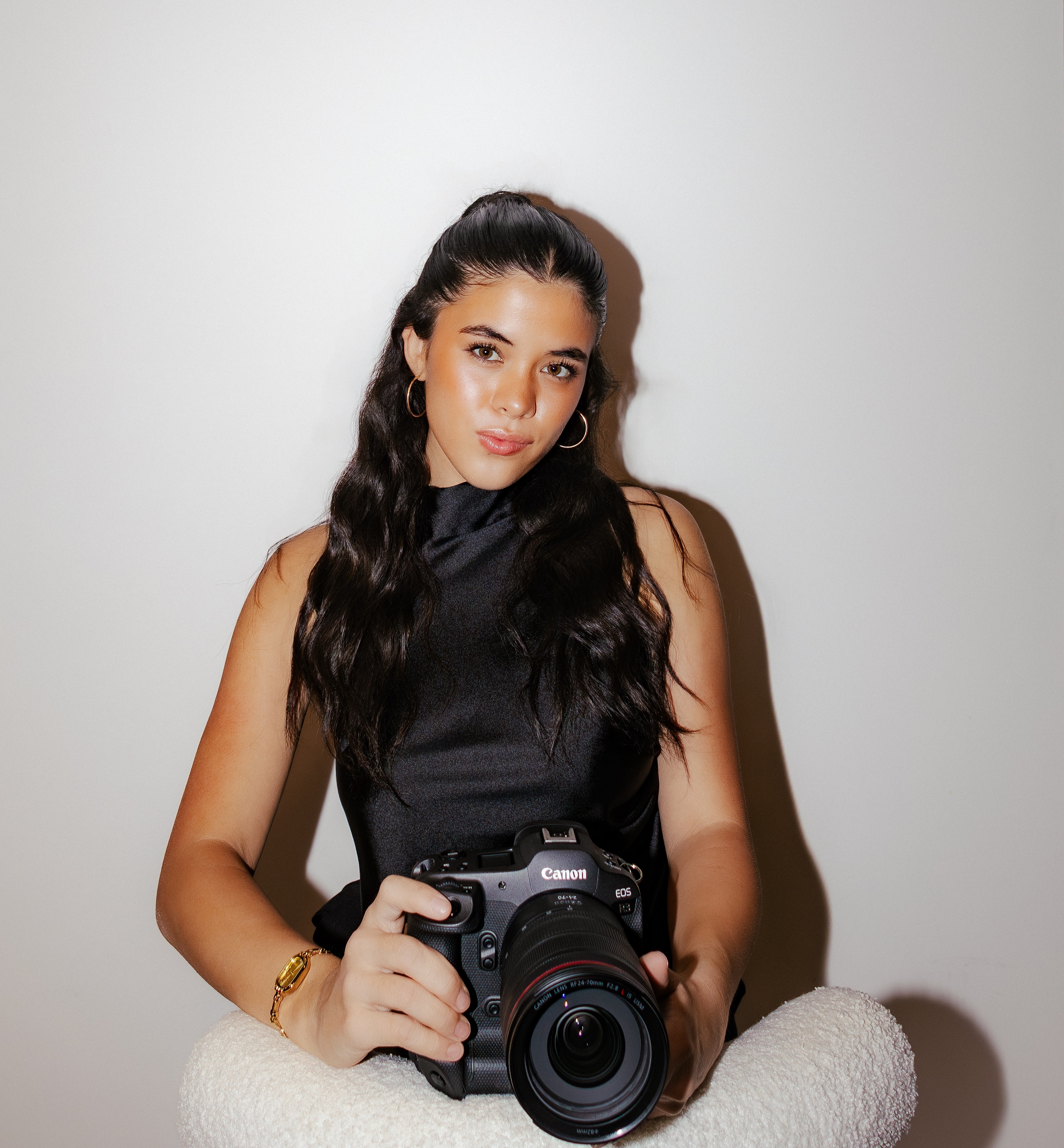 Woman with long dark hair holding a Canon camera, sitting against a white background.