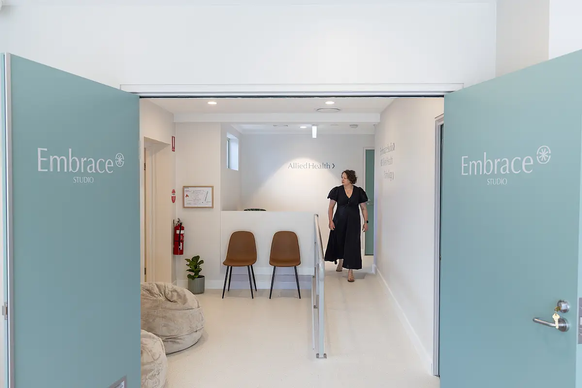 Open double doors with Embrace Studio logo leading to a bright hallway with two brown chairs, beige bean bags, and a woman in a black dress walking.