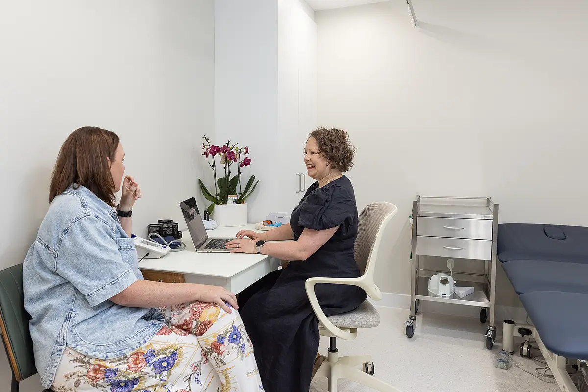 A healthcare professional smiling and typing on a laptop while consulting with a woman in a floral skirt and denim jacket in a bright medical office.