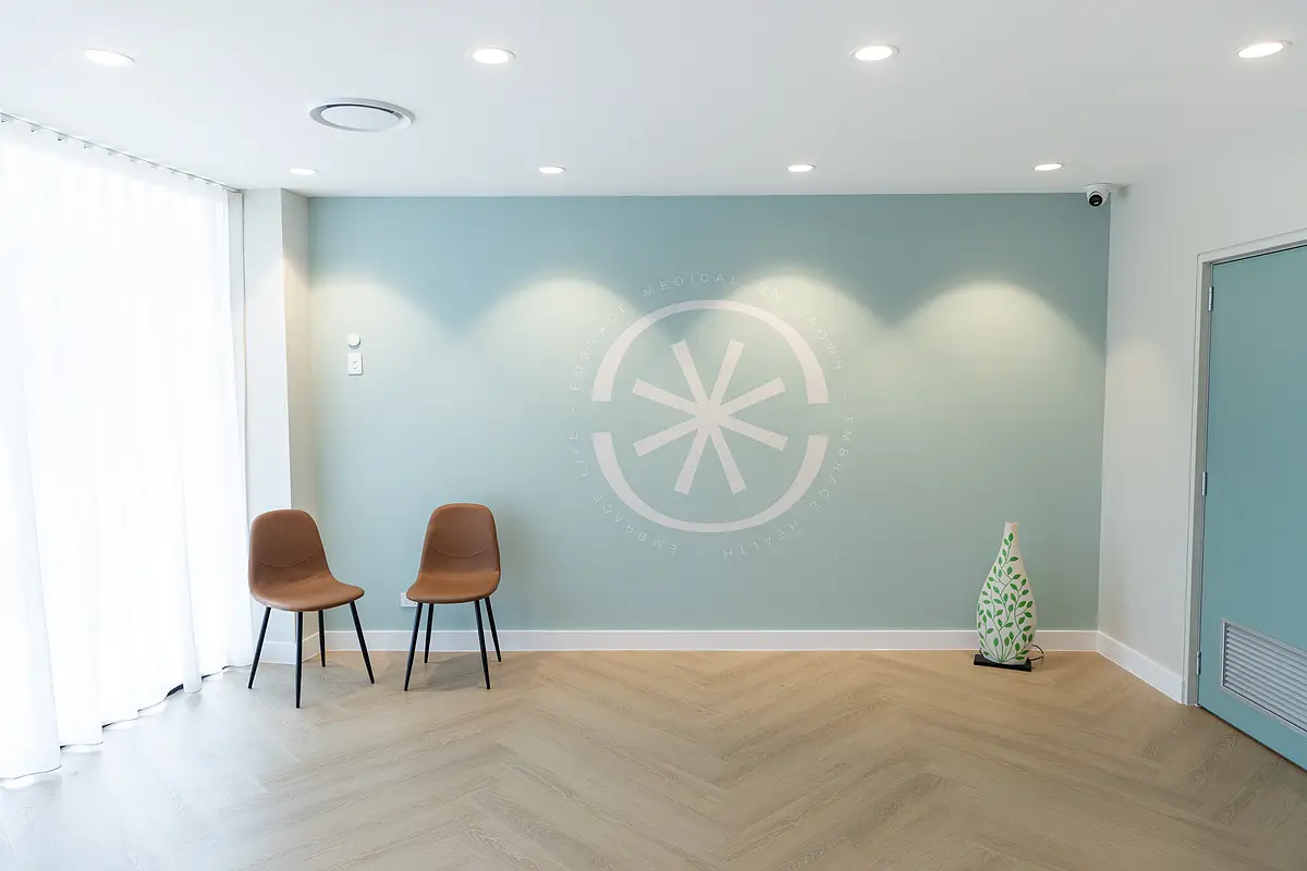 Minimalist room with two brown chairs, a light blue wall featuring a circular emblem, sheer curtains on the left, and a decorative vase on the floor to the right.