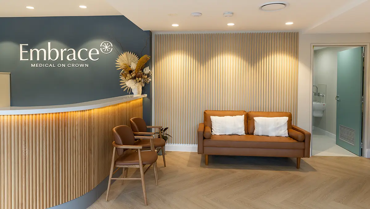 Modern medical office reception area with a curved wood panel desk, two brown chairs, a brown sofa with white pillows, and decorative dried plants.