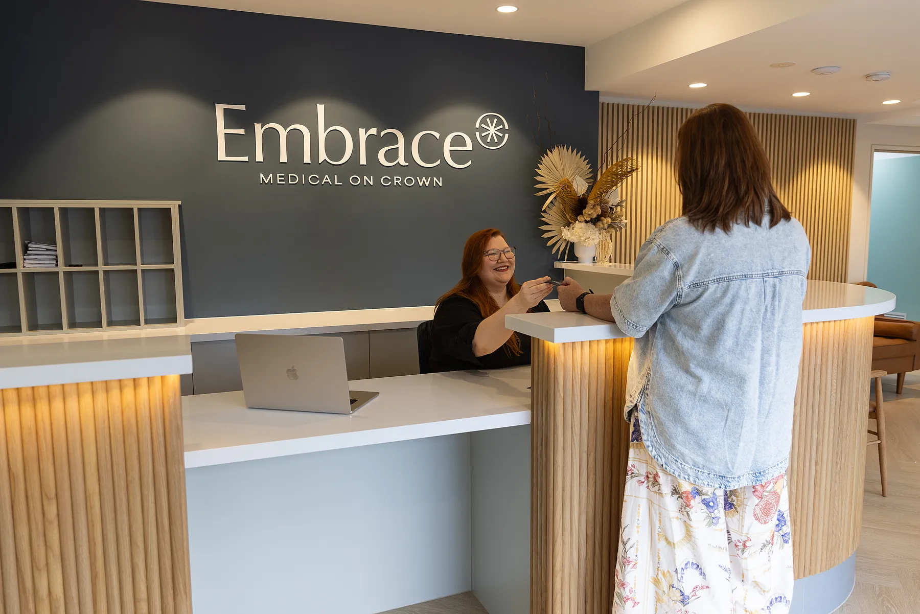 Receptionist smiling and handing a card to a visitor at the Embrace Medical on Crown reception desk.