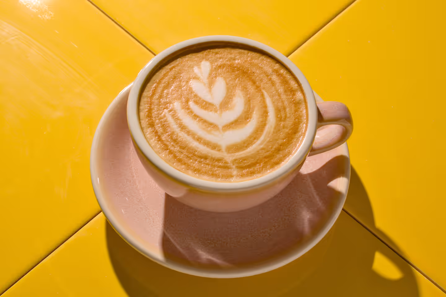 Cup of latte with leaf-shaped latte art on a pink saucer on a yellow tiled table.