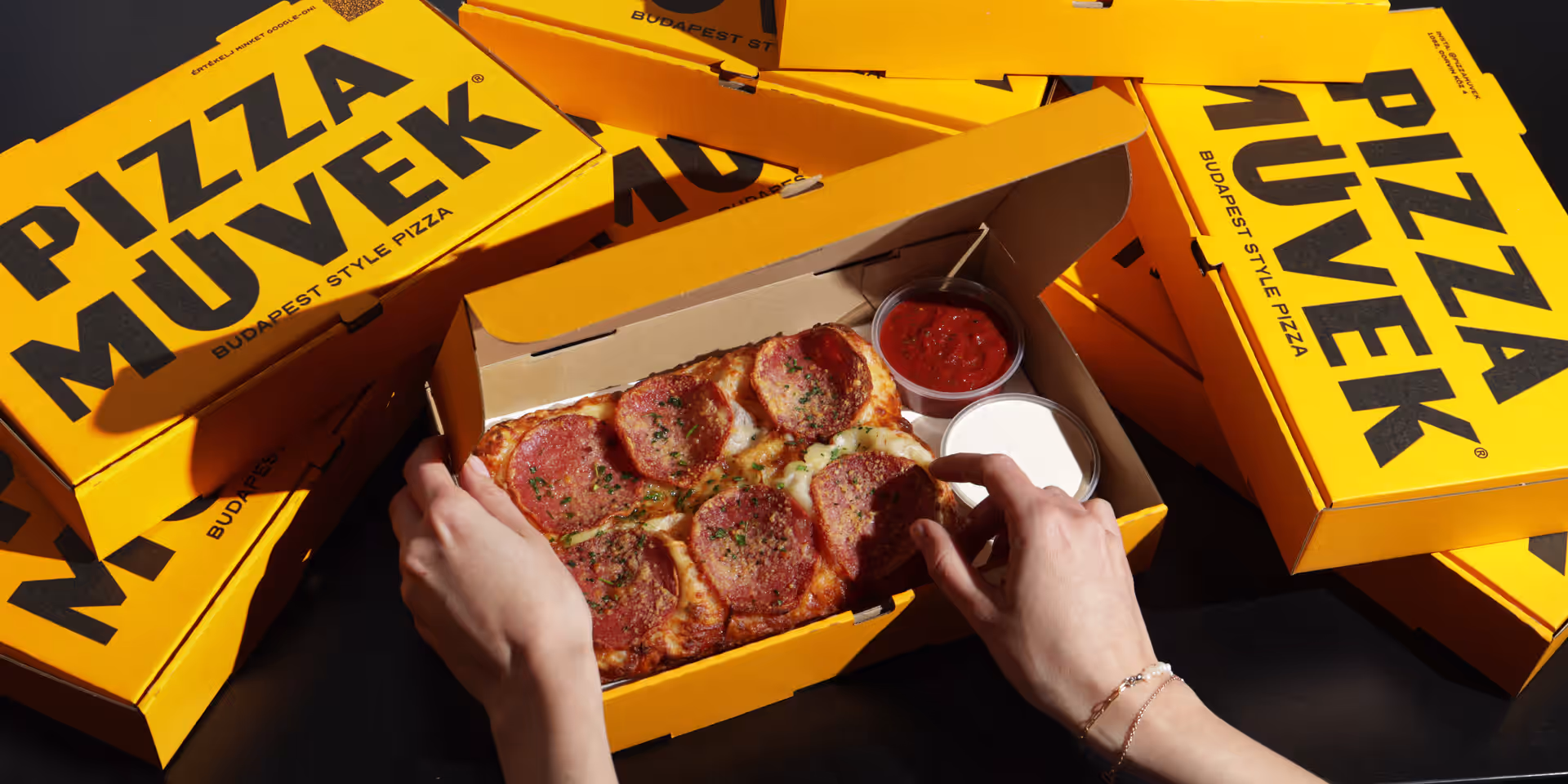 Hands holding open a box of budapest style salami pizza with red sauce and white dip in yellow Pizza Művek boxes.