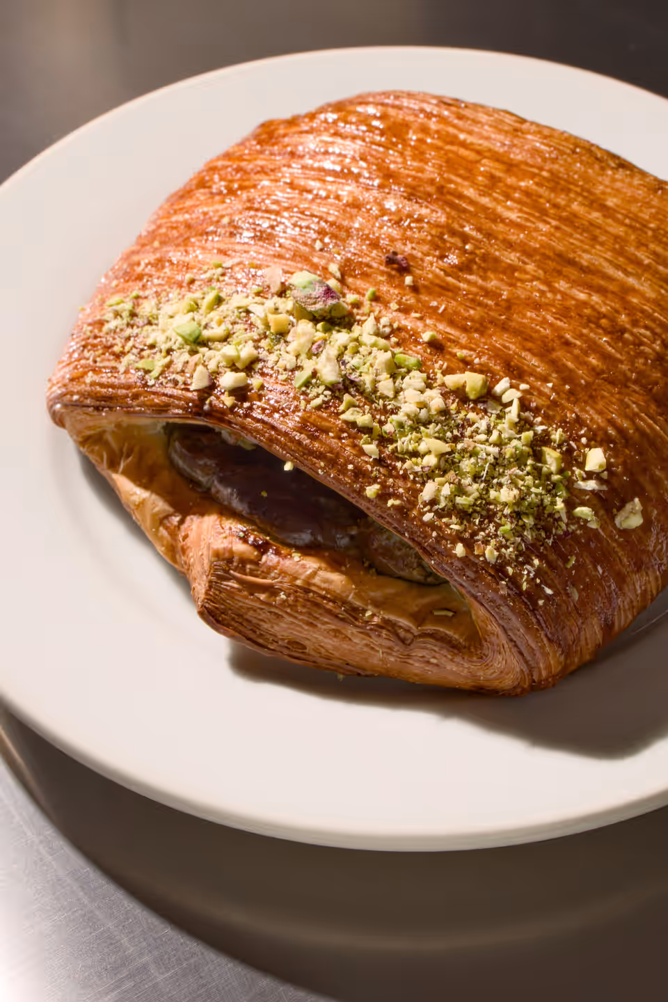 Flaky rectangular pastry topped with crushed pistachios on a white plate under warm light.