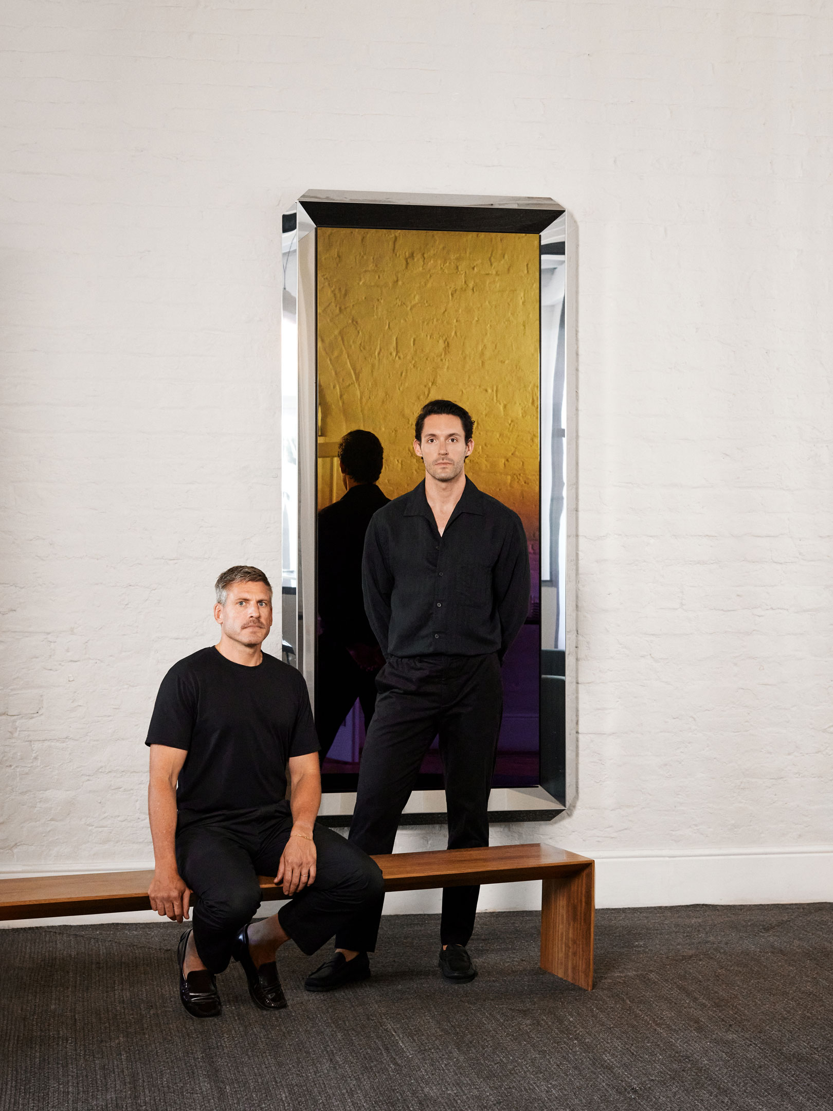 Two men dressed in black, one sitting on a wooden bench and the other standing behind it in front of a large mirror reflecting a yellow textured wall.