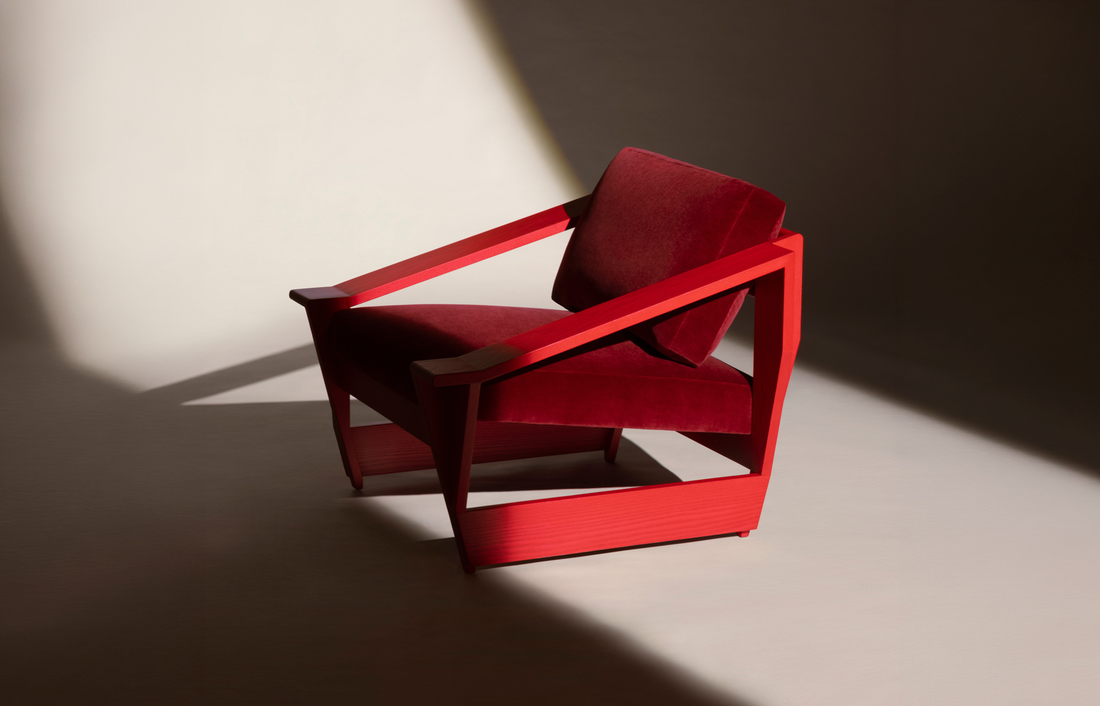 Modern chair with deep red cushion and bright red angular wooden frame placed on a light floor with dramatic shadows.