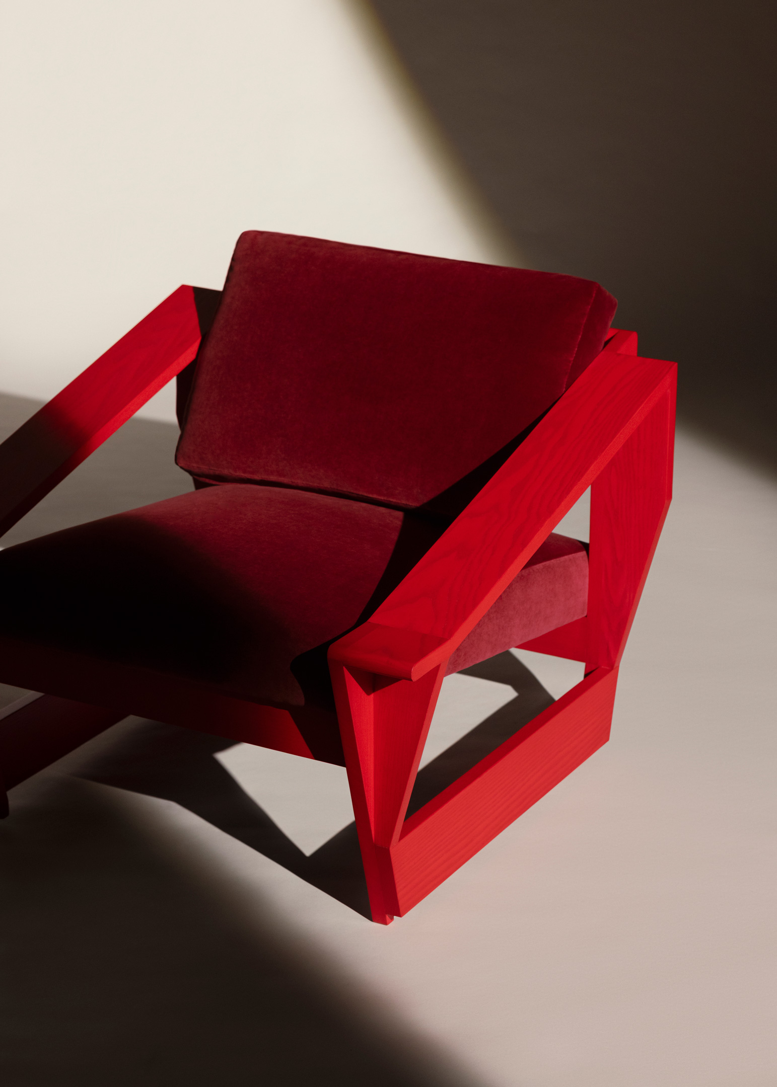 Modern armchair with a bright red wooden frame and deep red velvet cushions, partially illuminated by light on a neutral background.