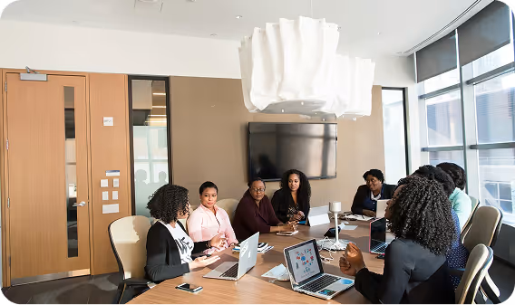 A diverse group of seven professional women seated around a conference table in a modern office, engaged in a meeting with laptops and notebooks.
