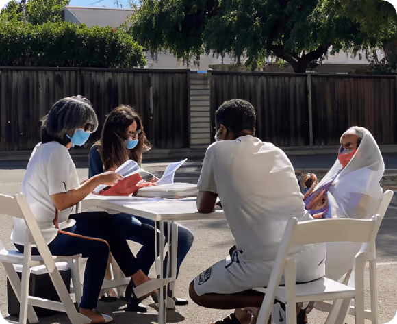 Four people wearing face masks sitting outdoors around a white table, engaged in reading and discussion.