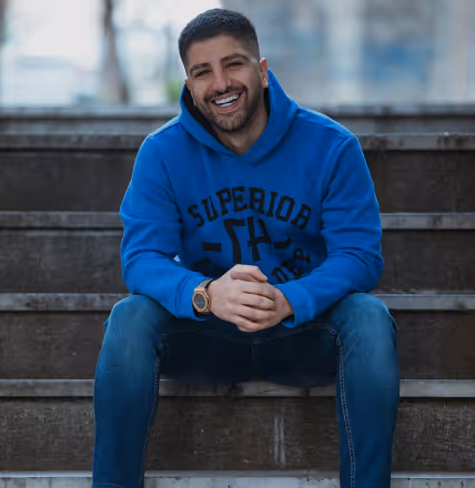 Smiling man in a blue hoodie and jeans sitting on outdoor steps.