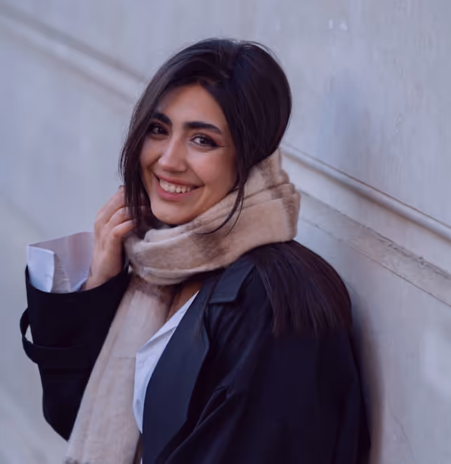 Smiling woman with long dark hair wearing a beige scarf and black coat leaning against a wall.
