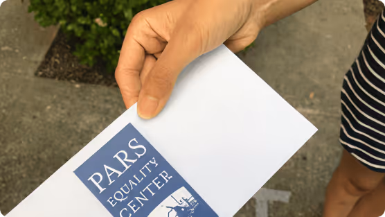 Hand holding an envelope with a blue Pars Equality Center logo outdoors.
