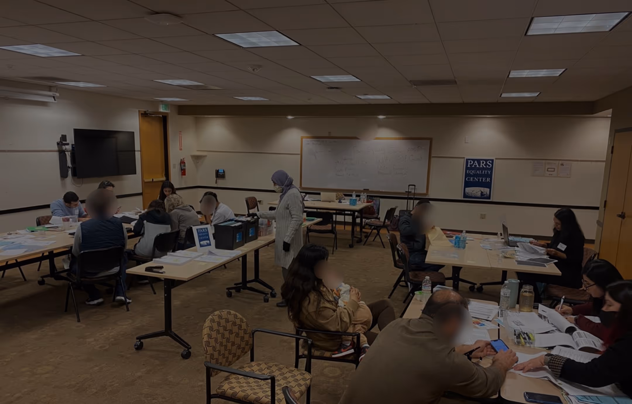 People seated at tables in a meeting room working on documents and using laptops, with a person standing wearing a hijab and mask.