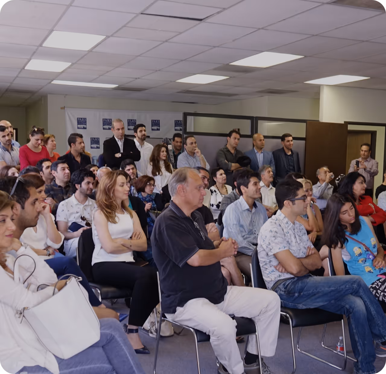 A diverse group of adults attentively sitting and standing in a room during a presentation or meeting.