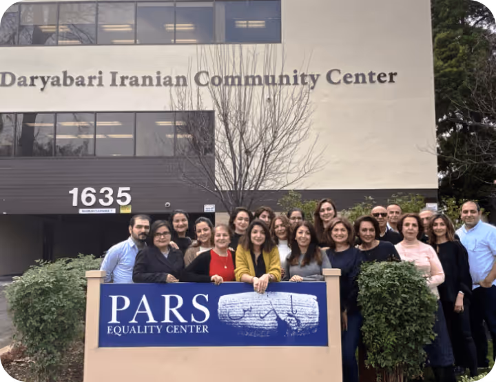 Group of people standing behind a sign for Pars Equality Center in front of the Daryabari Iranian Community Center building at 1635.